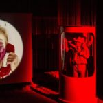 Zdjęcie przedstawia kadr ze spektaklu "Dzikość serca". Po lewej widoczny jest ekran na którym wyświetla się portret kobiety rozmawiającej przez telefon. której twarz ubrudzona jest różową szminką. Po prawej znajduje się oświetlona na czerwono budka, a wniej budka, a wniej widać kilka osób.