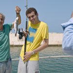 Kadr z filmu dwóch mężczyzn stoi na łodzi, jeden z nich trzyma wędkę, drugi prezentuje złowioną przez nich rybę