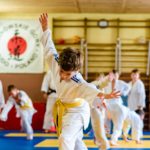 Zdjęcie przedstawia dzieci w tradycyjnych strojach judo podczas ćwiczeń ruchowych na sali gimnastycznej