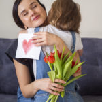 Zdjęcie przedstawia dziewczynkę przytulającą się do mamy, która w ręku trzyma bukiet czerwnych tulipanów i kartkę z ręcznie narysowanym serduszkiem