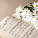 Zdjęcie przedstawia zbliżenie na rozłożony zeszyt z nutami i leżący na nim bukiet białych kwiatów