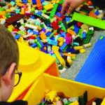 Zdjęcie przedstawia pudla pełne klocków LEGO i bawiące się nimi dzieci