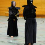Zdjęcie przedstawia dwie postaci w czarnym stroju kendo na sali gimnastycznej