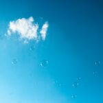 Fotografia przedstawia błękitne niebo, mały biały obłok i lecące bańki mydlane