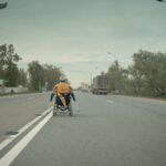 na wózku pokazany niepełnosprawny człowiek, który jedzie na wózku pośrodku drogi, obok niego jadą ciężarówki