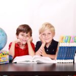 na zdjęciu dzieci siedzące za stołem, obok nich narzędzia szkolne: książki, zeszyty, globus